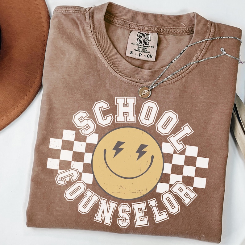 May include: A brown t-shirt with the words "SCHOOL COUNSELOR" in white block letters. A yellow smiley face with lightning bolt eyes is in the center, set against a white and black checkered background. A silver necklace with a small pendant rests on the shirt.