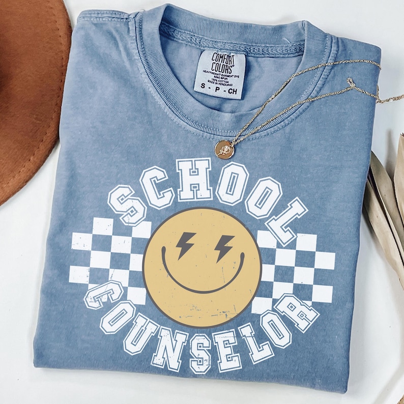 May include: A blue Comfort Colors t-shirt with the words "SCHOOL COUNSELOR" in white block letters. The design includes a yellow smiley face with lightning bolt eyes and a checkered background. A gold necklace with a pendant rests on the shirt.