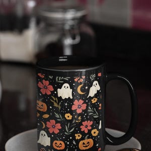 May include: Black ceramic mug with a Halloween-themed floral pattern. The design includes orange pumpkins, white ghosts, red flowers, and a crescent moon on a black background. The mug is on a gray saucer.