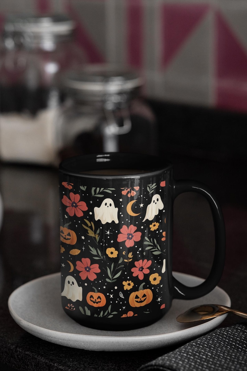 May include: Black ceramic mug with a Halloween-themed floral pattern. The design includes orange pumpkins, white ghosts, red flowers, and a crescent moon on a black background. The mug is on a gray saucer.