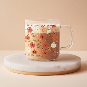 May include: A clear glass mug filled with a beverage, decorated with a Halloween-themed pattern. The design includes orange pumpkins, white ghosts, red flowers, and a crescent moon. The mug sits on a white and gold round coaster.