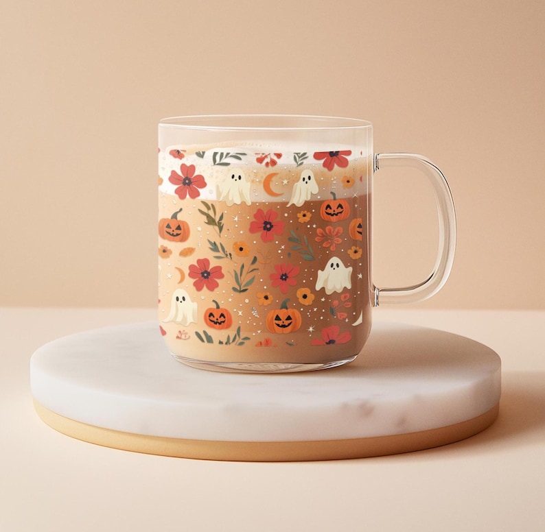 May include: A clear glass mug filled with a beverage, decorated with a Halloween-themed pattern. The design includes orange pumpkins, white ghosts, red flowers, and a crescent moon. The mug sits on a white and gold round coaster.
