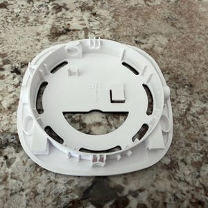 May include: A white plastic circular mounting bracket for a smoke detector or similar device. The component features a central opening, several curved slots, and a small moulded arrow pointing upward with the text UP printed next to it.