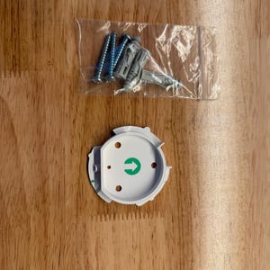 May include: A white mounting bracket with a green arrow and several screw holes, along with a clear plastic bag containing screws and wall anchors. The items are on a wooden surface.