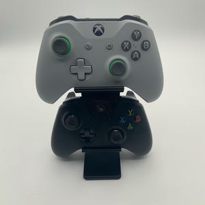 Puede incluir: Dos mandos de Xbox, uno gris y otro negro, apilados uno encima del otro. El mando gris tiene detalles en verde alrededor de los joysticks y los botones. El mando negro descansa sobre un soporte negro.