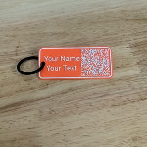 Könnte beinhalten: Orangener rechteckiger Schlüsselanhänger mit dem weißen Text "Your Name" und "Your Text". Auf der rechten Seite befindet sich ein QR-Code. Ein schwarzer Metallring ist an der linken Seite befestigt.