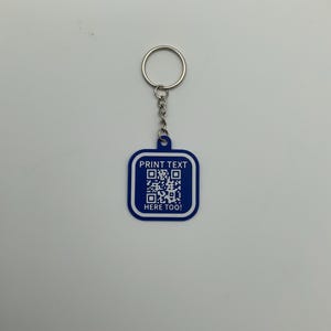 May include: A blue and white keychain with a QR code and the text "PRINT TEXT HERE TOO!". The keychain has a silver metal ring and chain. The design is on a square-shaped tag.