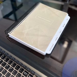May include: A beige home office journal with a black spiral binding and white tabs labelled "Meetings", "Reminders", "Notes", "To Do", and "Ideas". The journal has a textured, geometric pattern.