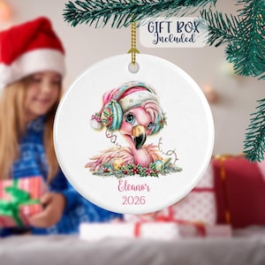 Può includere: Un ornamento rotondo in ceramica bianca con una colorata illustrazione di un fenicottero rosa che indossa un cappello da Babbo Natale e paraorecchie color ottanio. Il fenicottero è circondato da decorazioni natalizie. Il nome Eleanor e 2026 sono stampati.