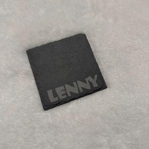 May include: A square, dark grey slate coaster with the name "LENNY" etched in a lighter grey font. The coaster is set against a soft, light grey background.