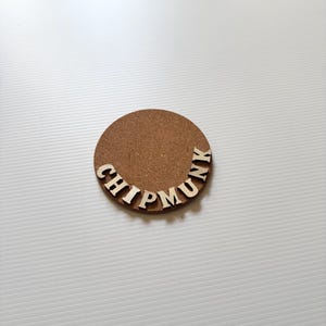 May include: A circular brown cork coaster featuring the word CHIPMUNK spelled out in raised, light-colored wooden capital letters arranged in a curved arc along the bottom edge of the coaster surface, creating a distinct textured appearance.