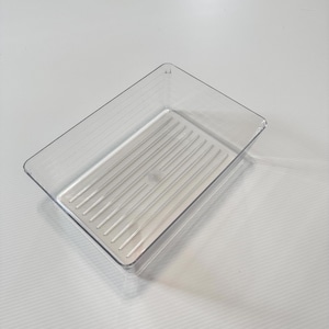 May include: A rectangular, clear plastic storage tray featuring a series of parallel, raised vertical ridges along the interior base. The container has smooth, transparent walls and an open top, designed for organizing small items or supplies.