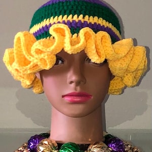 May include: A handmade crochet hat with a yellow, green, and purple color scheme. The hat features a ruffled brim and is displayed on a mannequin head. The hat is part of a Mardi Gras theme.