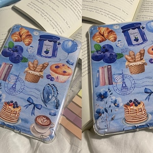 May include: A blue and white iPad case with a watercolor design featuring blueberries, croissants, butterflies, a stack of pancakes, a cup of coffee, and a stamp that says "Paris, France".