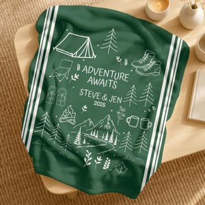 May include: A dark green blanket with white stripes and a camping design. The blanket features illustrations of a tent, trees, hiking boots, and the text "ADVENTURE AWAITS STEVE & JEN 2025".