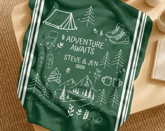 May include: A dark green blanket with white stripes and a camping-themed design. The blanket features illustrations of a tent, trees, hiking boots, and the text "ADVENTURE AWAITS STEVE & JEN 2025".