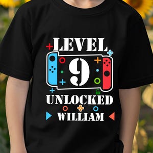 May include: A black t-shirt with the text "LEVEL 9 UNLOCKED WILLIAM" and a graphic of a game controller. The shirt is being worn by a young person in front of a background of sunflowers.