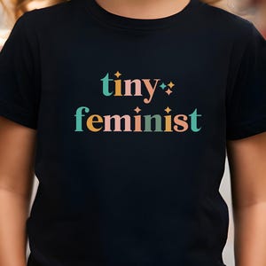 May include: A black t-shirt with the words "tiny feminist" in colorful text. The shirt is worn by a young person with long, wavy hair and blue earrings. The background is blurred, suggesting an outdoor setting.