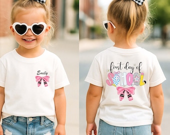 Könnte beinhalten: Weißes T-Shirt mit rosa Schleife und Ballett-Schuh-Design. Das Shirt hat den Namen "Emily" über der Schleife aufgedruckt. Auf der Rückseite des Shirts steht "first day of school" mit bunten Buchstaben und einer passenden Schleife und Ballettschuhen.
