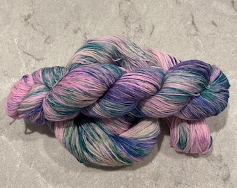 May include: A skein of multi-coloured yarn in pink, purple, teal, and cream. The yarn is twisted into a large, knotted bundle, displaying the colour variations. The yarn appears soft and suitable for knitting or crochet projects.
