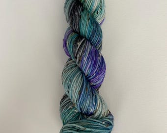 Puede incluir: Un ovillo de hilo en tonos de verde azulado, morado y gris con motas blancas. El hilo está retorcido en forma de espiral.