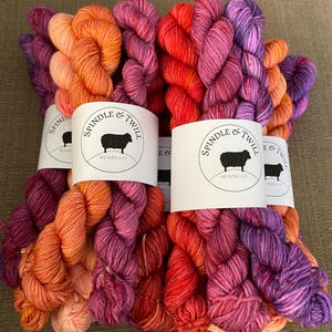 Könnte beinhalten: Mehrere Stränge farbenfroher Wolle in Orange-, Rot-, Rosa- und Lilatönen. Jeder Strang ist mit einem weißen Etikett mit der Aufschrift "SPINDLE & TWILL WEAVING CO" und einem Schaf-Logo versehen. Die Wolle ist ansprechend angeordnet.