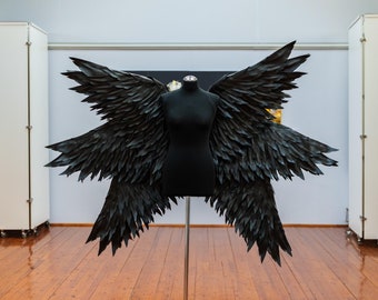 May include: A black mannequin with large, black, feathered wings on a stand in a room with wooden floors and white walls.