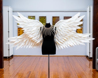 May include: A black dress form with large white paper angel wings. The wings are layered and detailed. A gold-colored collar is around the neck. The dress form is on a metal stand, set on a wooden floor.
