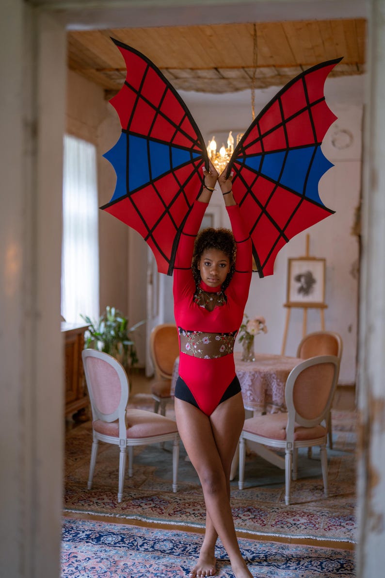 May include: A red bodysuit with black accents and a floral mesh panel. The model is holding up large, stylized bat wings in red, black, and blue. The wings have a web-like design.