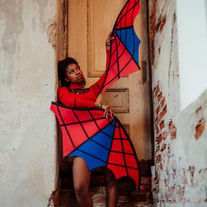 May include: A red long-sleeved top and matching skirt with a geometric design in red, blue, and black. The outfit is worn in a doorway with a distressed wall and brick. The outfit resembles wings.