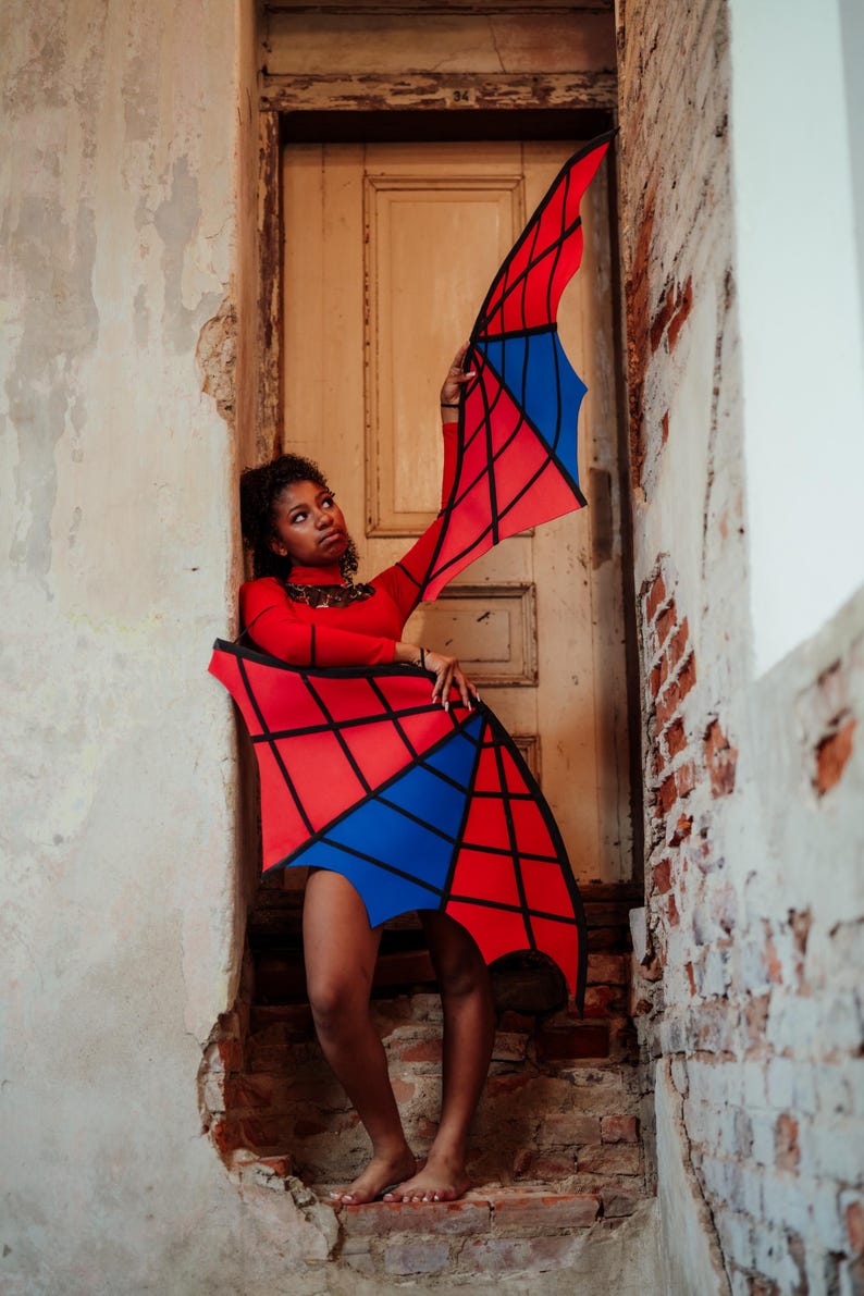 May include: A red long-sleeved top and matching skirt with a geometric design in red, blue, and black. The outfit is worn in a doorway with a distressed wall and brick. The outfit resembles wings.