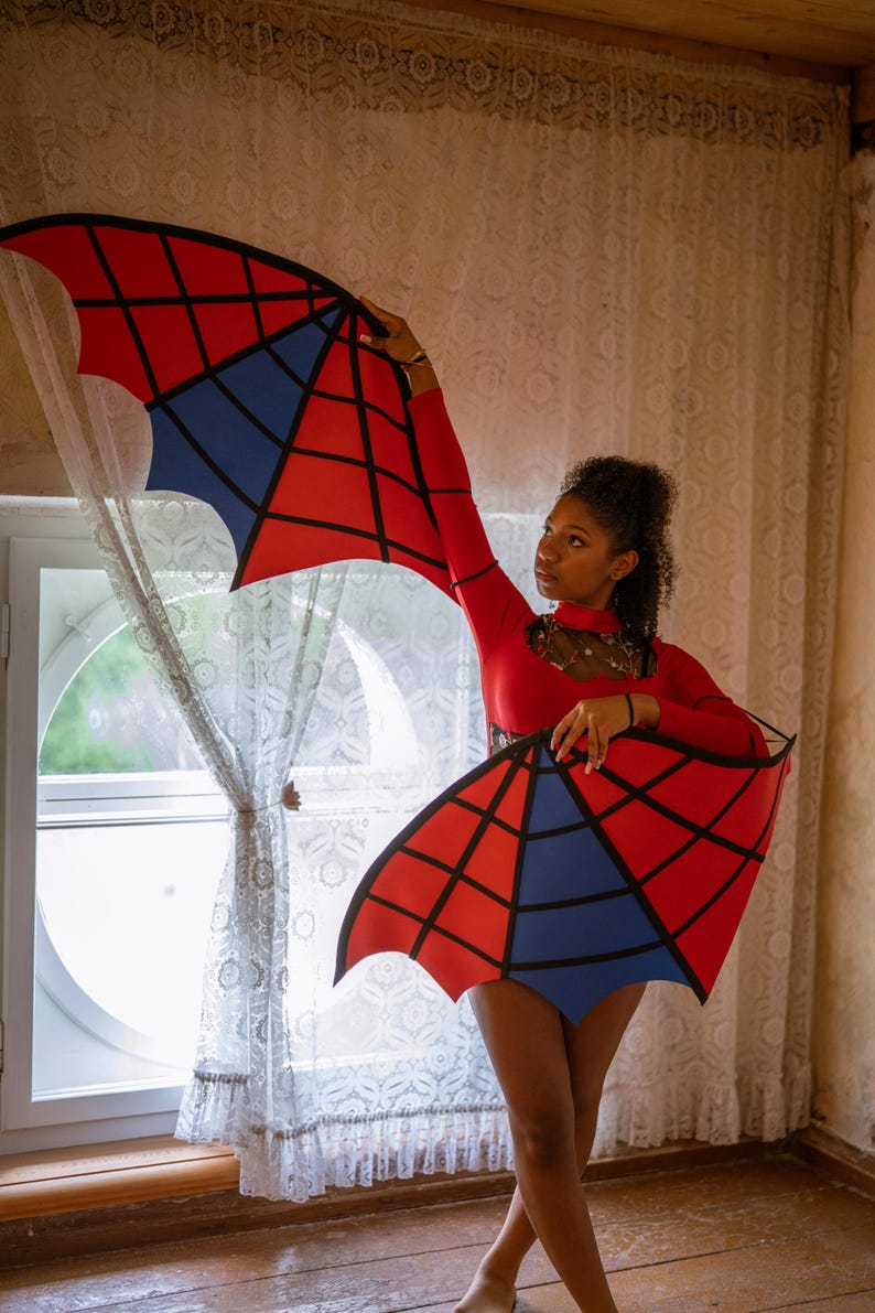 May include: A person wearing a red bodysuit with matching red and blue geometric wings. The wings have a black outline and a spiderweb design. The outfit is worn in front of a window with lace curtains.