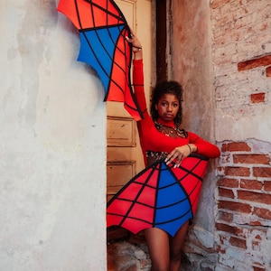 May include: A red, blue, and black costume with wing-like structures. The wings are red and blue with black lines, and the costume is worn with a red long-sleeve top. The costume is displayed in a doorway with a brick wall.