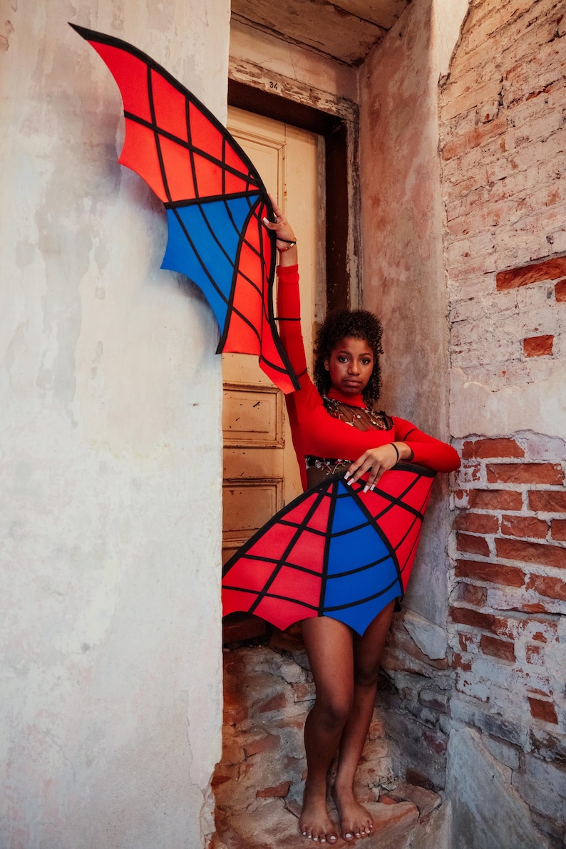 May include: A red, blue, and black costume with wing-like structures. The wings are red and blue with black lines, and the costume is worn with a red long-sleeve top. The costume is displayed in a doorway with a brick wall.
