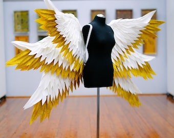 May include: A pair of large white and gold angel wings on a black mannequin stand. The wings are made of feathers and have a glittery finish.