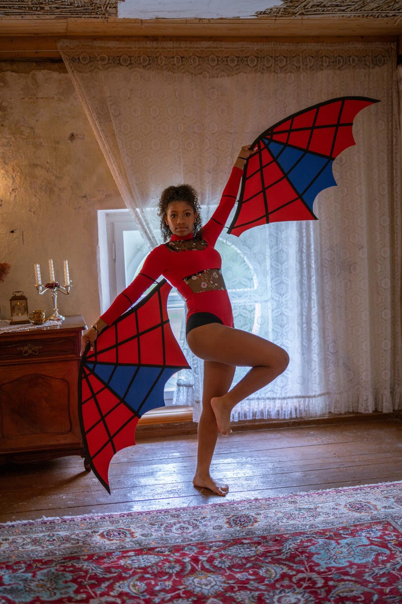 May include: A red bodysuit with long sleeves and a black bottom. The person is holding two large, stylized wings with a red and blue geometric design. The wings are in the shape of a bat's wings. The background includes a window, curtains, and a wooden cabinet.