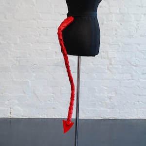 May include: A long, flexible red tail accessory constructed from overlapping scale-like segments. The tail features a pointed, triangular arrow-shaped tip at the end and is attached to a black waistband, displayed on a black mannequin torso.
