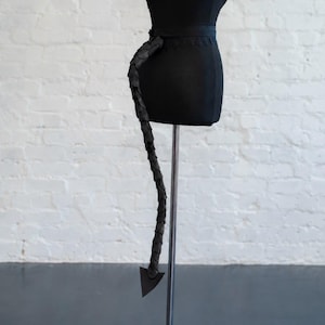 Può includere: Un busto di manichino nero indossa una cintura nera con una lunga coda segmentata composta da pezzi sovrapposti a forma di scaglie e una punta triangolare. La cintura è decorata con un bordo in pizzo lungo il bordo superiore.