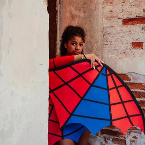 May include: A red long-sleeved top and blue shorts are paired with a geometric red and blue wing-like accessory. The accessory has a black outline and is held against a brick wall.