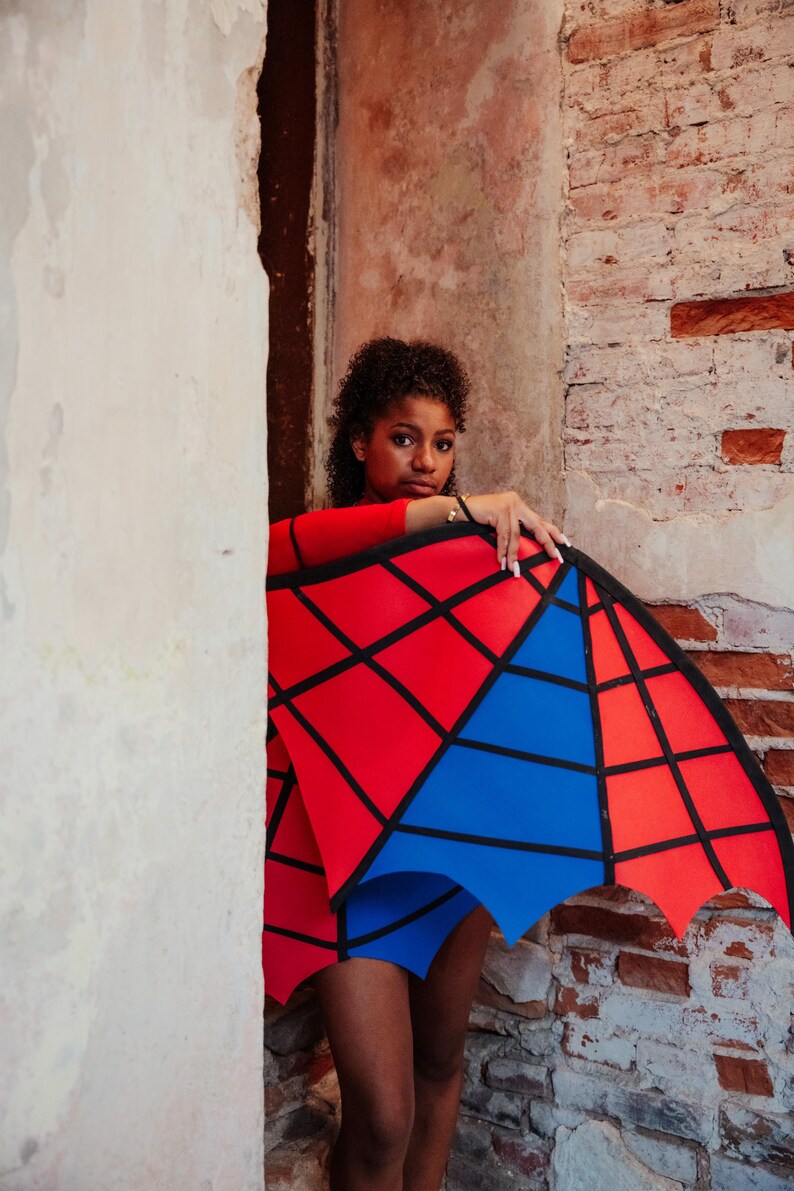 May include: A red long-sleeved top and blue shorts are paired with a geometric red and blue wing-like accessory. The accessory has a black outline and is held against a brick wall.