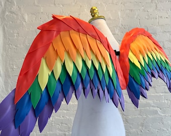 May include: A pair of large, decorative wings featuring layered, pointed feathers in a vibrant rainbow gradient of red, orange, yellow, green, blue, and purple. The wings are displayed on a white mannequin torso, showcasing the structured design.