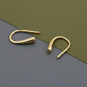 May include: A pair of gold-colored, curved earrings. Each earring has a smooth, rounded shape, with a hook-like design. The earrings are displayed on a two-tone background of gray and olive green.