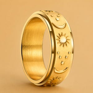May include: A gold-colored ring with a celestial design. The ring features embossed suns, crescent moons, and stars around the band. The ring has a smooth, polished finish and a rotating outer band.