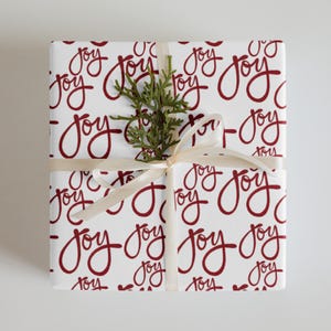 Könnte beinhalten: Verpacktes Geschenk mit weißem Papier mit dem Wort "Joy" in sich wiederholender burgunderfarbener Schrift. Ein Zweig Grün und ein cremefarbenes Band schmücken das Paket und schaffen eine festliche und elegante Präsentation.