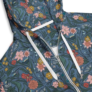Puede incluir: Una sudadera con capucha azul con cremallera y un estampado floral detallado de flores rosas, amarillas y naranjas, junto con hojas y tallos verdes. La sudadera tiene un cordón blanco y una cremallera plateada.