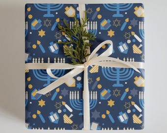 Puede incluir: Un regalo envuelto con papel azul marino con un patrón de menorás, dreidels, velas y estrellas en amarillo y blanco. Una ramita de vegetación y una cinta color crema completan la presentación.
