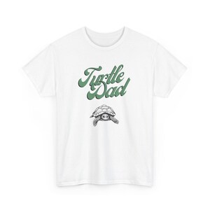 May include: White t-shirt with the text "Turtle Dad" in green script above a detailed illustration of a turtle. The design is centered on the shirt, offering a casual and nature-themed aesthetic.