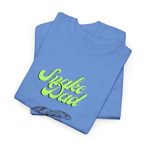 May include: Two light blue t-shirts are folded and stacked. The top shirt features the text "Snake Dad" in neon green with a black outline, and a detailed illustration of a snake's head.