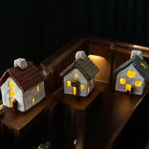 Pode incluir: Três casas em miniatura de cerâmica com janelas e portas iluminadas. As casas têm telhados vermelhos, verdes e azuis, chaminés e detalhes decorativos. Estão colocadas numa superfície de madeira.