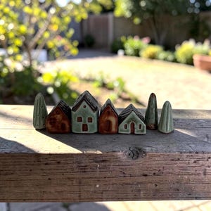 May include: A collection of miniature ceramic figurines featuring four small houses and three stylized trees. The houses are glazed in shades of brown and sage green with simple etched windows and doors. The trees are smooth, tapered, and green.
